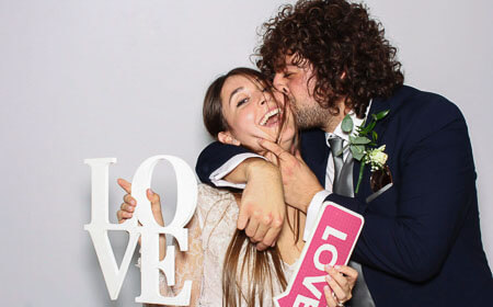 A groom kissing his bride in a photo booth while she holds 2 love signs